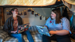 Two Honors students it on a dorm room bed together using their laptops, talking and smiling.
