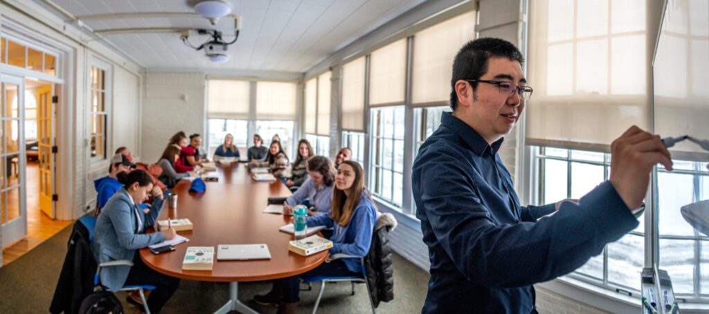 A group of 15 Honors students sit around a table in class in Balentine Hall, while their Preceptor Hao Hong stands at the front of the class writing on a white board.