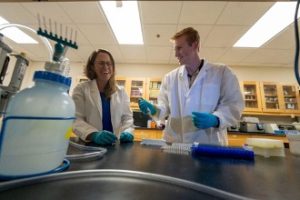 Two people in a science lab wearing lab coats and blue gloves 