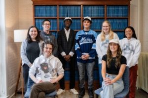 A group of students wearing UMaine clothing in front of the Thesis bookshelves