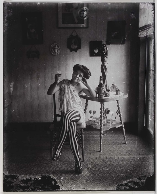 A black and white photo of a woman in striped stockings sitting at a table drinking something from a wine glass. 